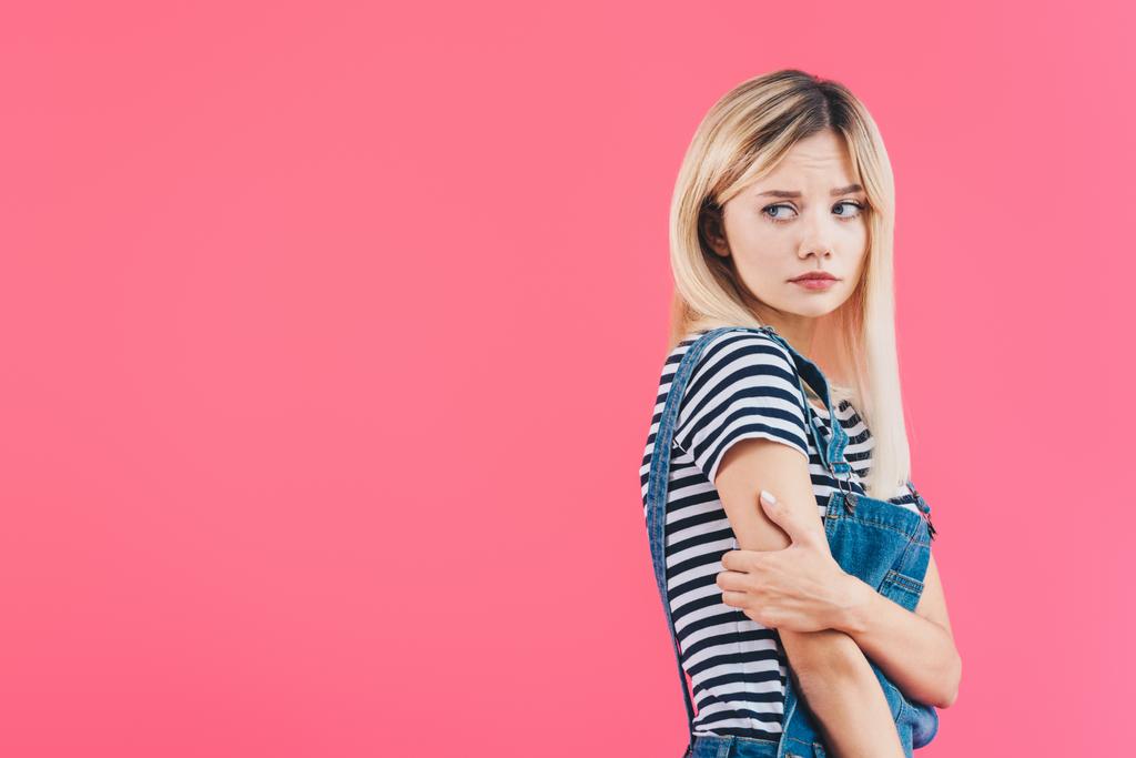 sad girl in denim overall hugging herself and looking away isolated on pink - Photo, Image