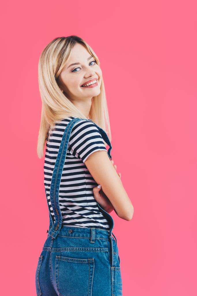 smiling beautiful girl in denim overall standing with crossed arms and looking at camera isolated on pink - Photo, Image