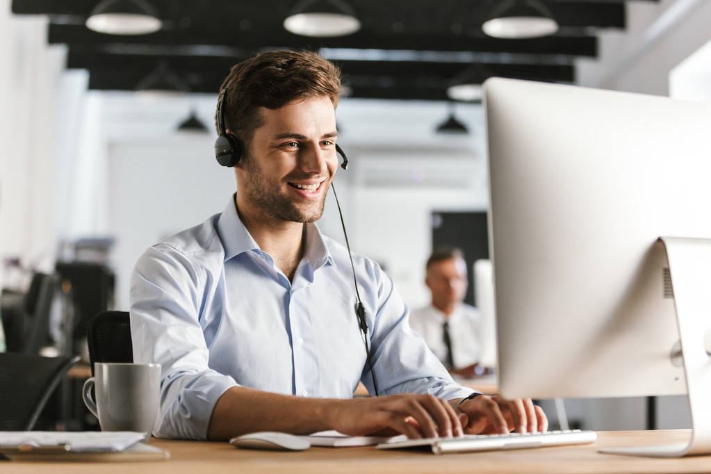 Zdjęcie z noszenia ubrania biurze sprawny człowiek 20s i zestaw słuchawkowy działa na komputerze w call center - Zdjęcie, obraz