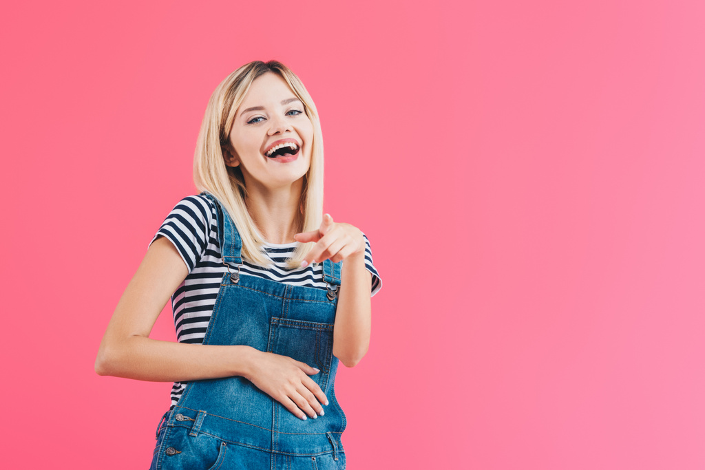 дівчина сміється в джинсовому режимі, вказуючи на камеру ізольовано на рожевому
 - Фото, зображення