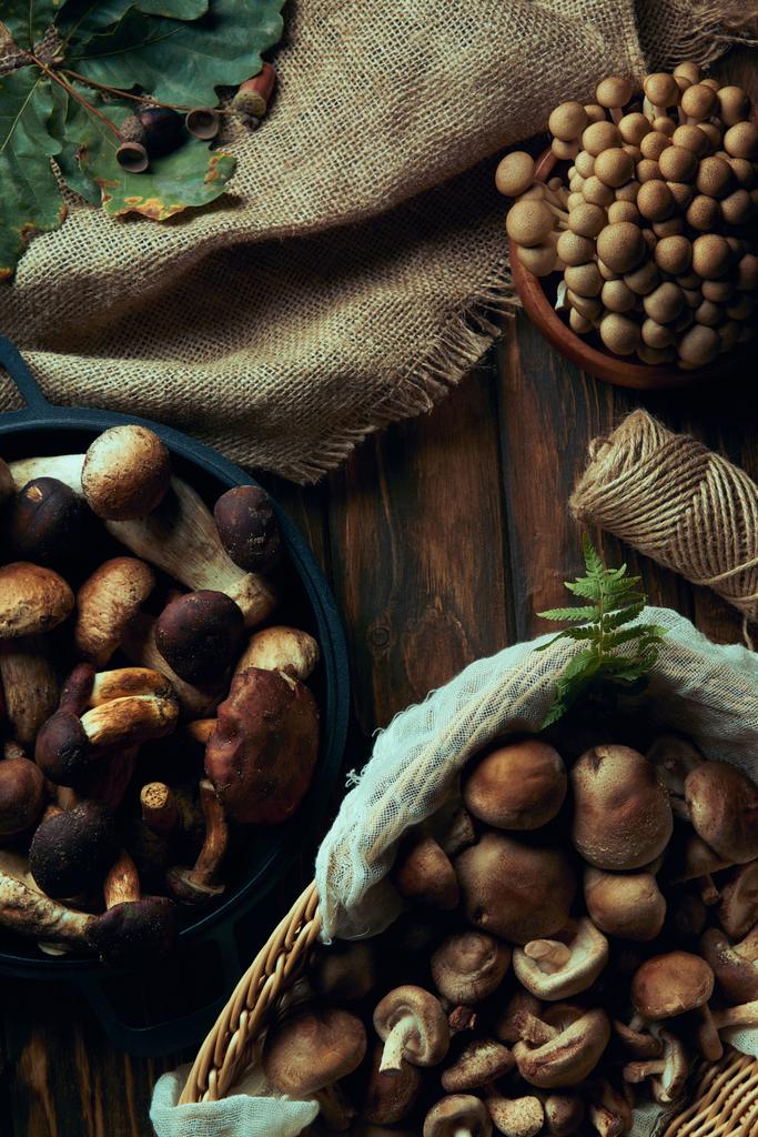 vista superior de cogumelos comestíveis crus variados em cesta e tigelas, pano de saco e corda na mesa de madeira
 - Foto, Imagem