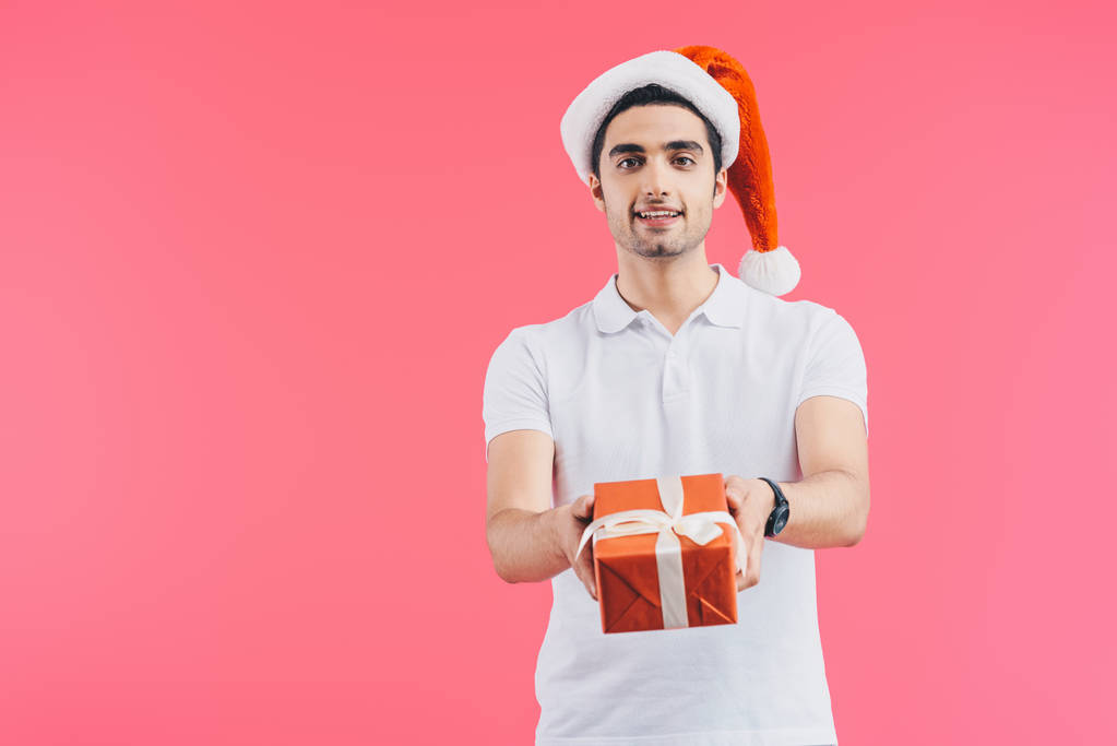 усміхнений красивий чоловік у капелюсі Санта дарує подарункову коробку ізольовано на рожевій, новорічній концепції
 - Фото, зображення