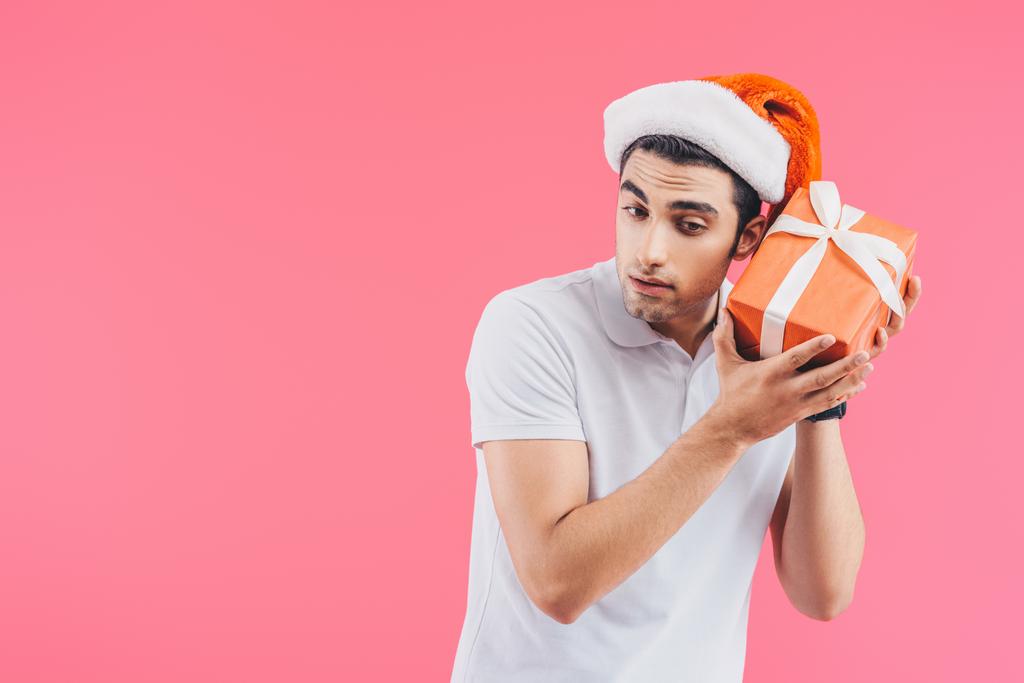 excited handsome man in santa hat listening gift box isolated on pink, new year concept - Photo, Image