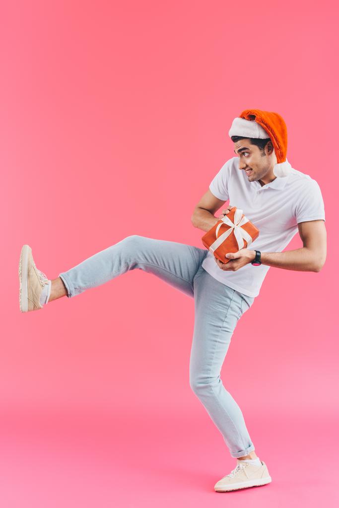 Grimassenhaft schöner Mann mit Weihnachtsmütze und erhobenem Bein hält Geschenkbox isoliert auf rosa, Neujahrskonzept - Foto, Bild