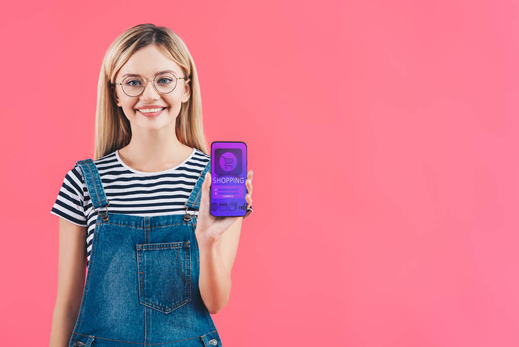 portrait of smiling woman in eyeglasses showing smartphone with shopping sign isolated on pink - Photo, Image