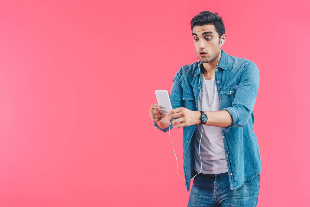 Porträt eines schockierten Mannes mit Kopfhörern, der in Händen isoliert auf rosa das Smartphone betrachtet - Foto, Bild