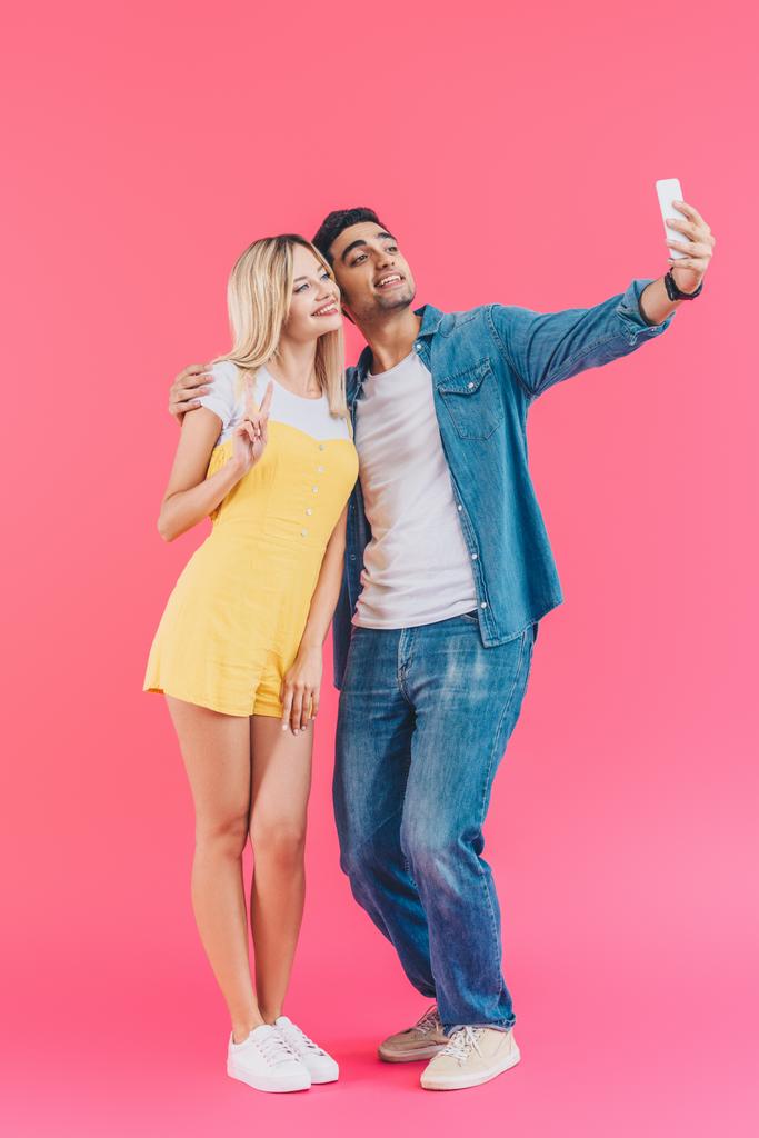 handsome young man taking selfie with girlfriend doing peace sign on smartphone isolated on pink  - Photo, Image