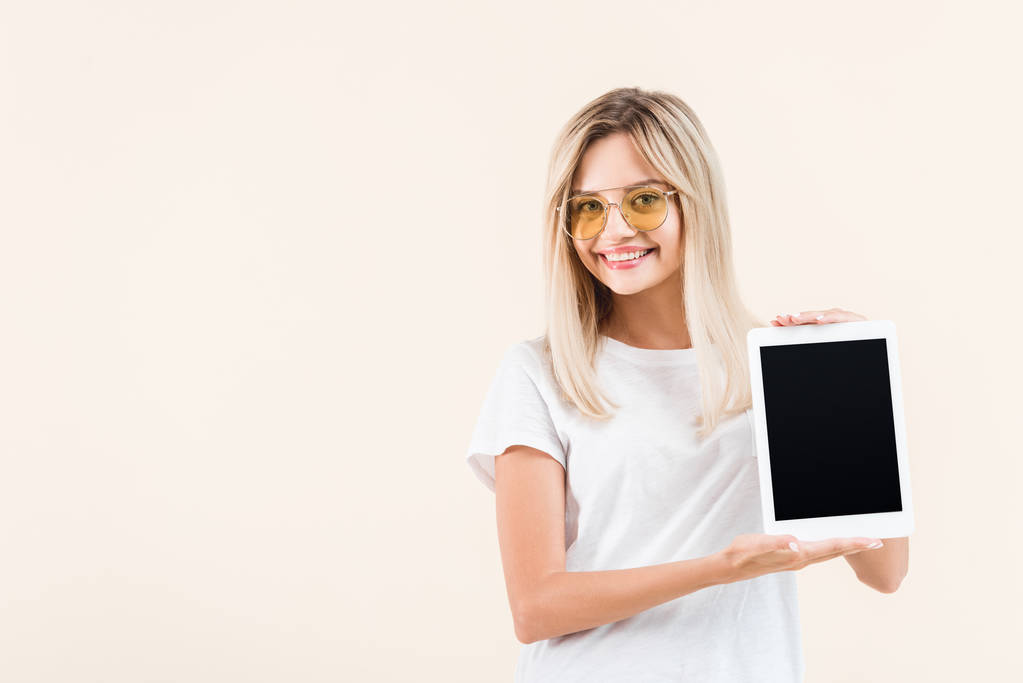 šťastná mladá žena v brýlích ukazuje digitální tabletu s prázdnou obrazovkou izolované na béžové - Fotografie, Obrázek