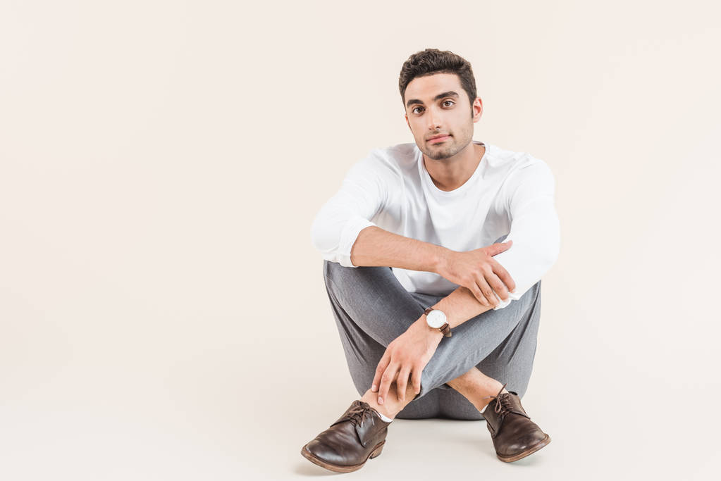 full length view of handsome young man sitting and looking at camera isolated on beige - Photo, Image