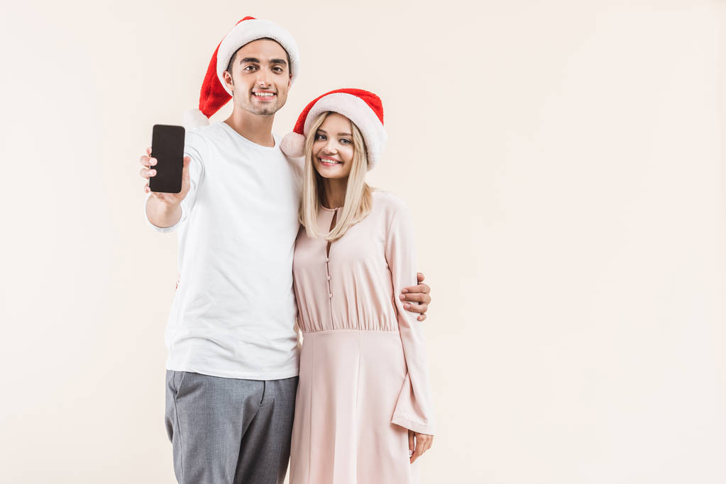 jovem casal em santa chapéus segurando smartphone com tela em branco e sorrindo para a câmera isolada no bege
  - Foto, Imagem