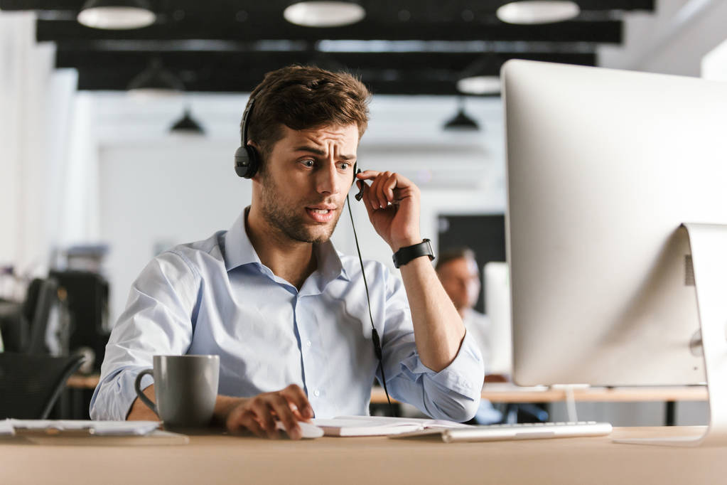 Kuva stressaantunut mies 30s yllään toimistovaatteet ja kuulokkeet istuu tietokoneella ja työskentelee call center
 - Valokuva, kuva