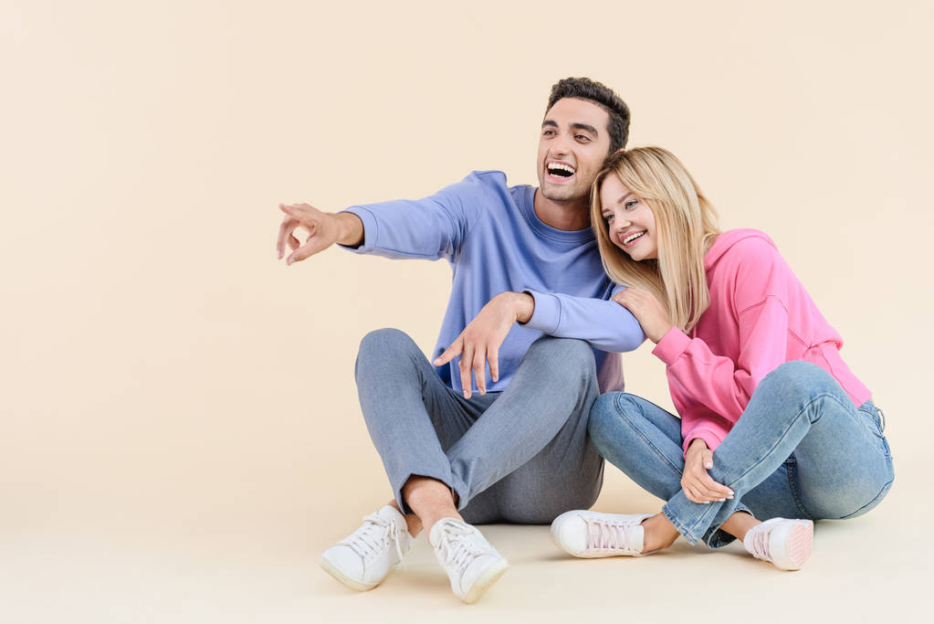 cheerful young couple sitting together and pointing away isolated on beige - Photo, Image