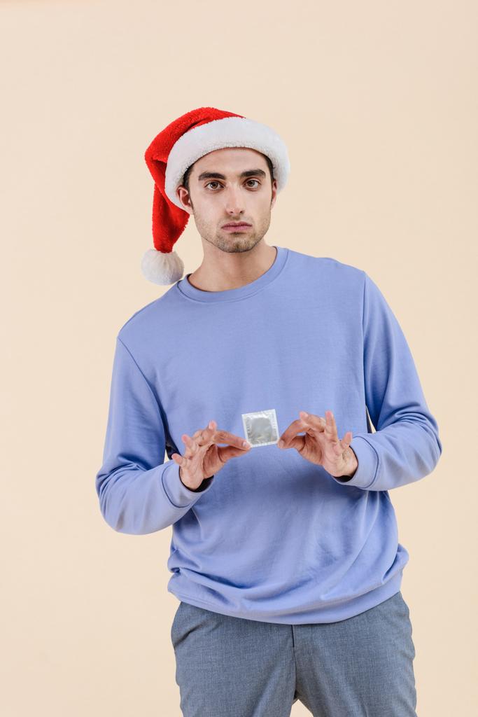 yakışıklı genç adam prezervatif tutarak ve bej izole kamera bakarak Noel Baba şapkası - Fotoğraf, Görsel