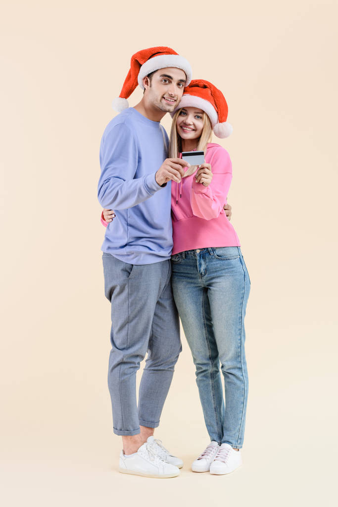 felice giovane coppia in cappelli di Babbo Natale in possesso di carta di credito e sorridente alla fotocamera isolata sul beige
 - Foto, immagini