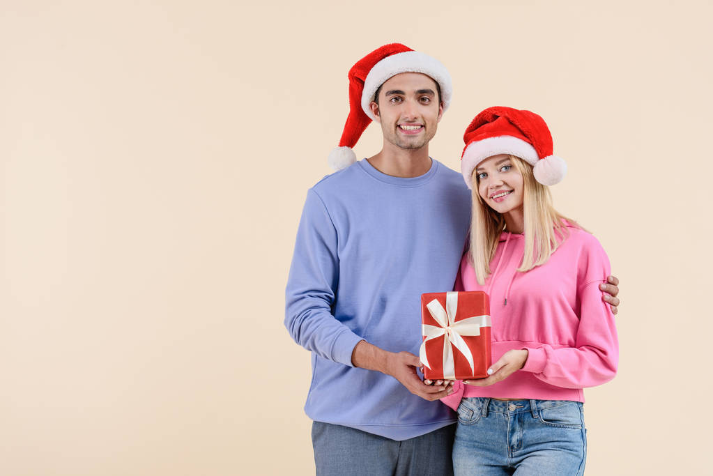 ευτυχισμένο ζευγάρι σε καπέλα santa κρατώντας χριστουγεννιάτικο δώρο και χαμογελαστός στην κάμερα που απομονώνονται σε μπεζ - Φωτογραφία, εικόνα