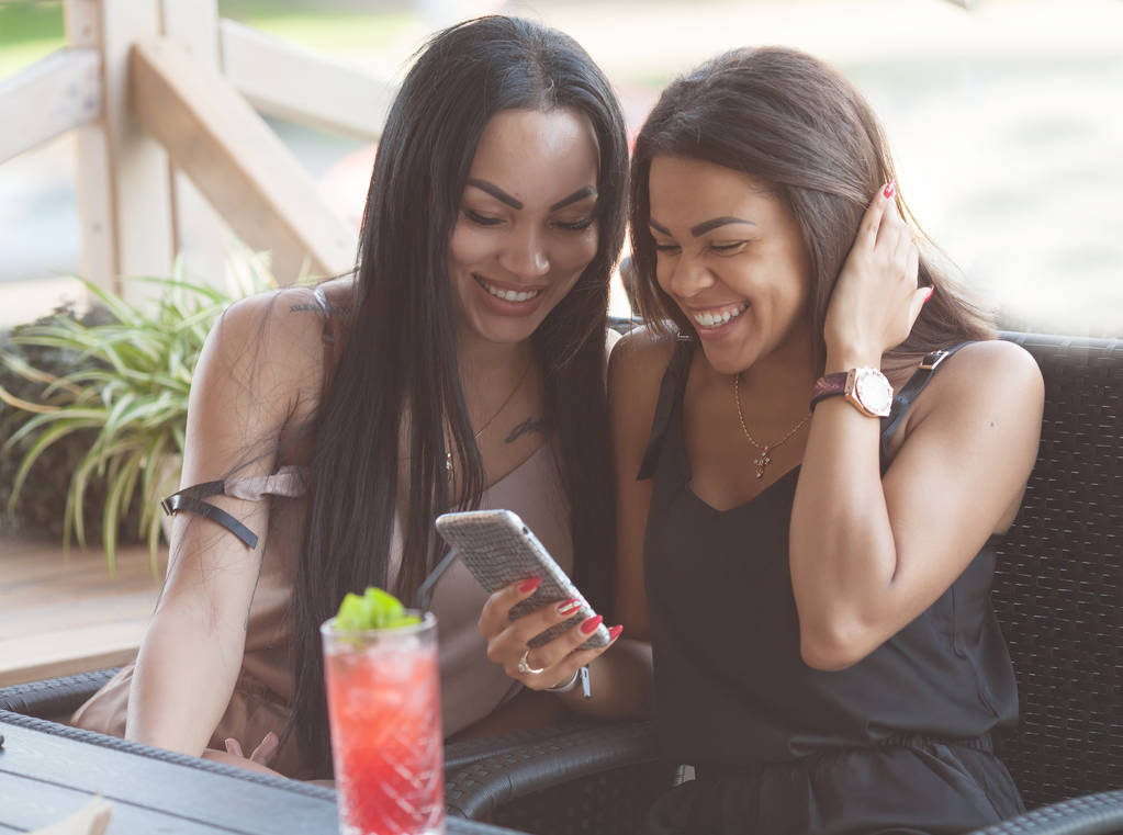 Multi-etnisch vrouwen lezen kennisgeving met positief nieuws op mobiele telefoon, vrije tijd in Terrascafé, omhelzen elkaar, hebben gelukkig expressies. Twee diveristy vrouwtjes kiezen iets in internet - Foto, afbeelding