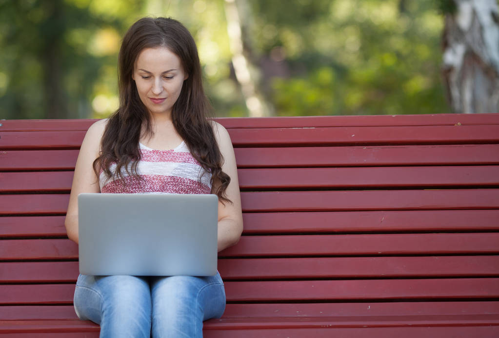 Beautiful caucasian woman is sitting at the bench in a city park with a laptop connected to wi-fi. Entrepreneur is working on a portable computer while sitting outdoors on a blurred urban background. - Photo, Image