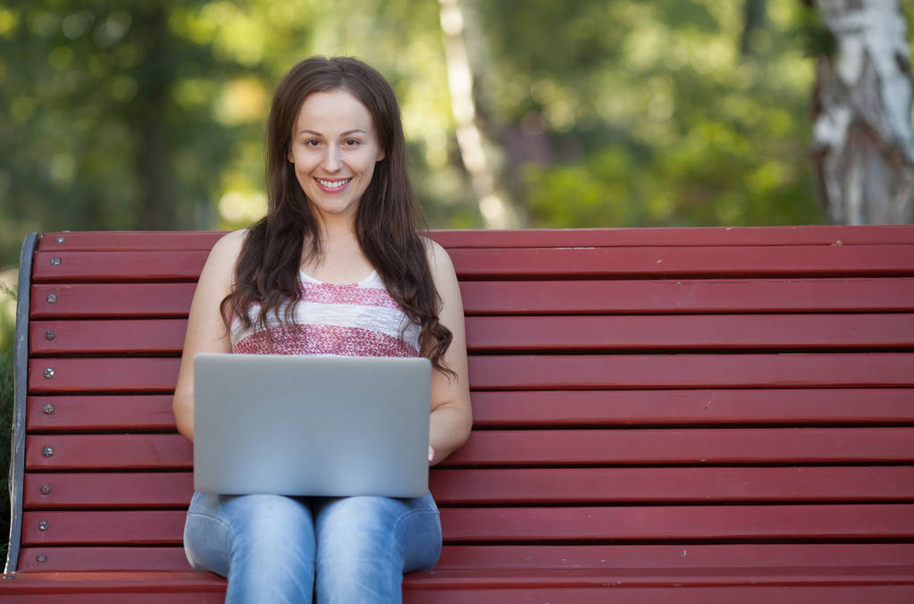 Красивая белая женщина сидит на скамейке в городском парке с ноутбуком, подключенным к wi-fi. Предприниматель работает за портативным компьютером, сидя на открытом воздухе на размытом городском фоне
. - Фото, изображение