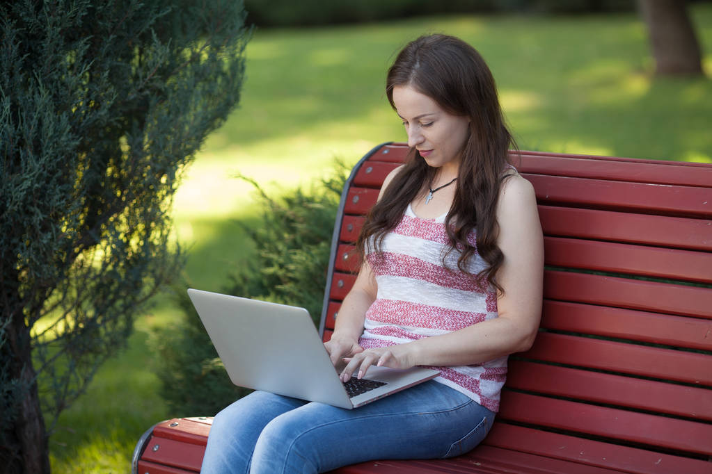 Krásná Kavkazský žena sedí na lavičce v městském parku s notebookem připojeni k wi-fi. Podnikatel pracuje na přenosném počítači při posezení venku na rozostřeného pozadí městské. - Fotografie, Obrázek