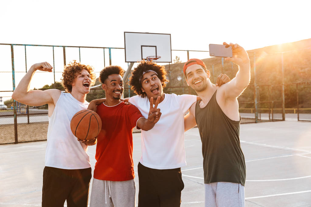 Foto de chicos alegres y deportivos sonriendo y tomando selfie en el teléfono inteligente mientras juega al baloncesto en el patio al aire libre durante el día soleado de verano
 - Foto, imagen