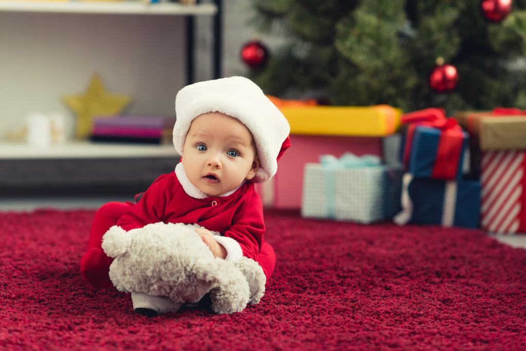 Close-up πορτρέτο της όμορφο μωράκι σε santa κοστούμι που βρίσκεται στο κόκκινο χαλί με αρκουδάκι μπροστά από το χριστουγεννιάτικο δέντρο και τα δώρα και να βλέπουν τα φωτογραφικών μηχανών - Φωτογραφία, εικόνα