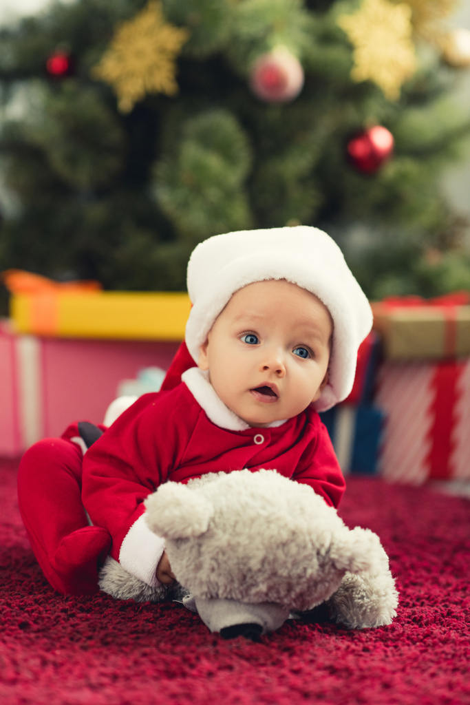 primer plano retrato de lindo bebé en traje de santa acostado en la alfombra roja con oso de peluche delante del árbol de Navidad y regalos
 - Foto, imagen