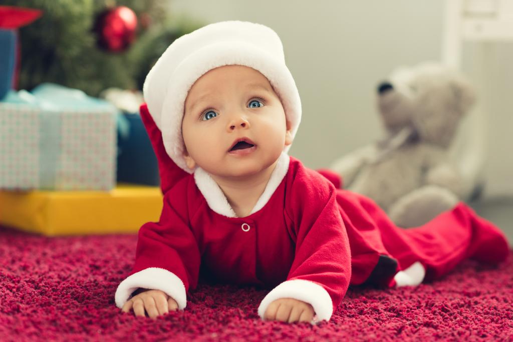 かわいい小さな赤ちゃんクリスマス プレゼントをして背景にぼやけとレッド カーペットに横たわってサンタ スーツで - 写真・画像