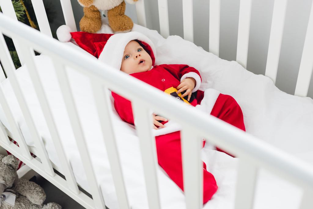 μεγάλη γωνία θέα της χαριτωμένο μικρό μωρό σε santa κοστούμι ξαπλωμένη στο παχνί με αρκουδάκι και βλέπουν τα φωτογραφικών μηχανών - Φωτογραφία, εικόνα