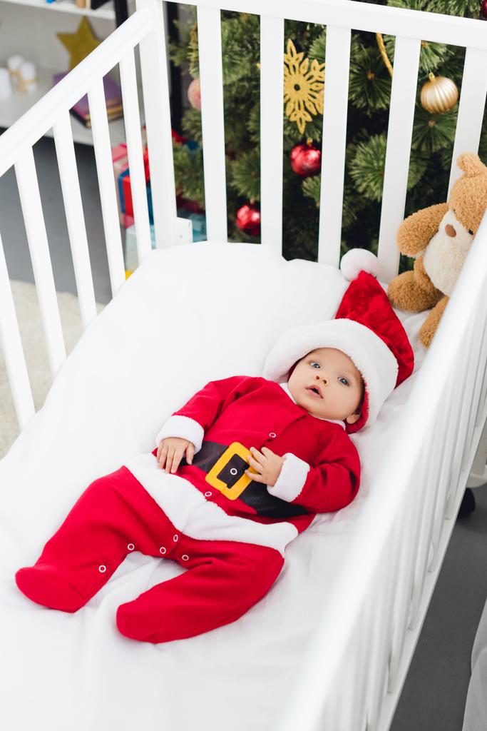 hoge hoekmening van mooie kleine baby in santa pak liggend in bedje met teddybeer en camera kijken - Foto, afbeelding