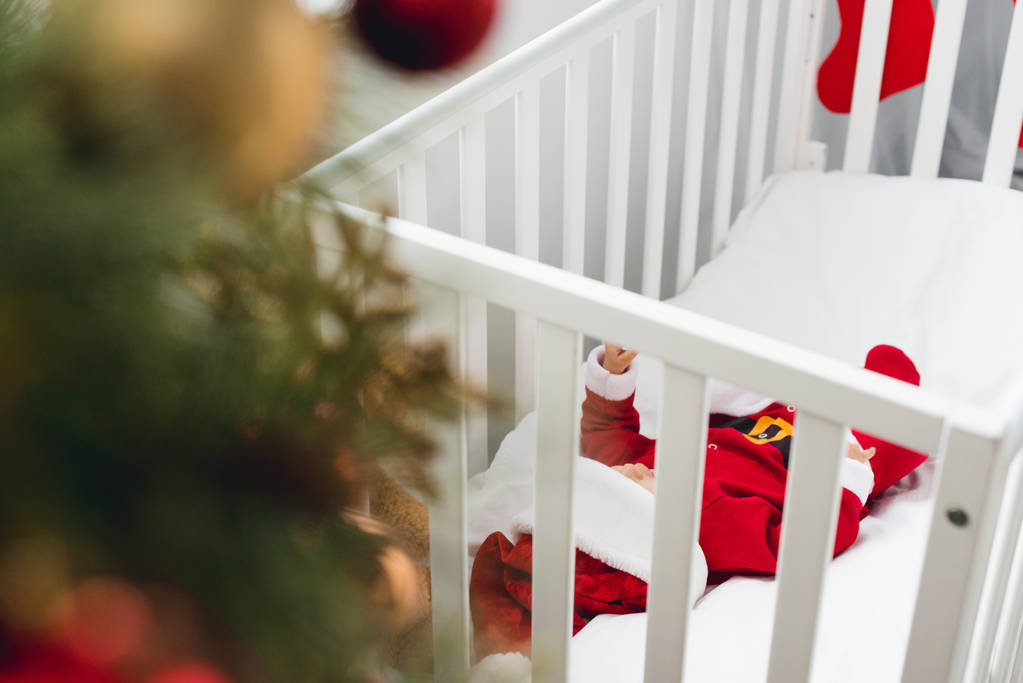 schattige kleine baby in santa pak liggend in bedje met kerstboom op voorgrond - Foto, afbeelding