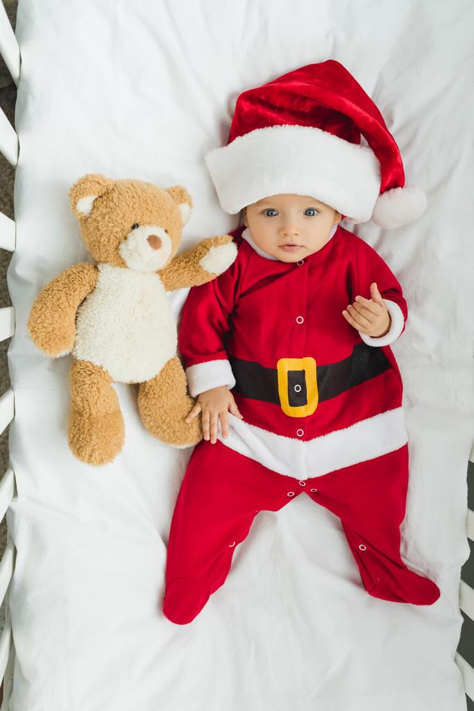 bovenaanzicht van schattige kleine baby in santa pak liggend in bedje met teddybeer en camera kijken - Foto, afbeelding