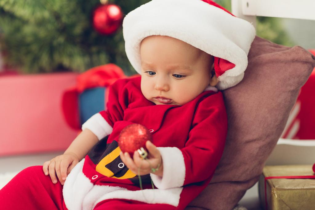entzückendes kleines Baby im Weihnachtsanzug mit Christbaumkugel - Foto, Bild