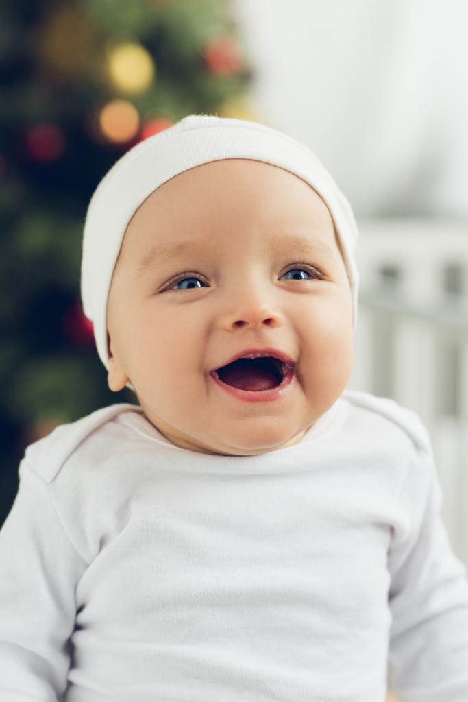 Nahaufnahme Porträt eines lachenden kleinen Babys mit weißem Hut und verschwommenem Weihnachtsbaum auf dem Hintergrund - Foto, Bild