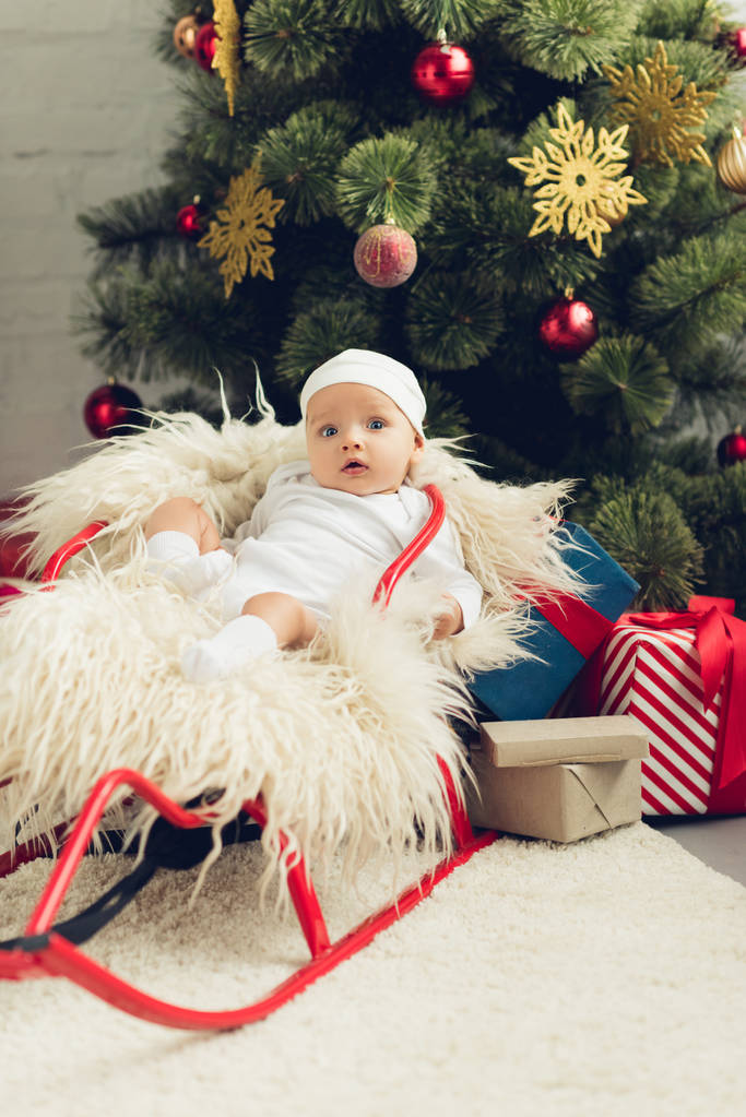 entzückendes kleines Baby im Schlitten vor dem Weihnachtsbaum mit Geschenken - Foto, Bild