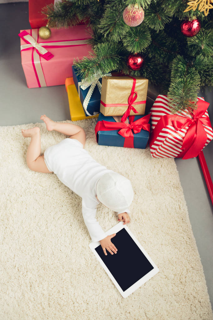 vista de ángulo alto de bebé pequeño con la tableta que miente en el piso cerca de regalos de Navidad y árbol
 - Foto, Imagen