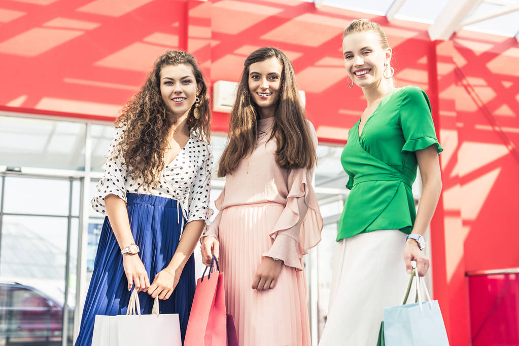 nízký úhel pohledu krásné stylové mladých žen drží papírové tašky a usmívá se na kameru v nákupní centrum  - Fotografie, Obrázek