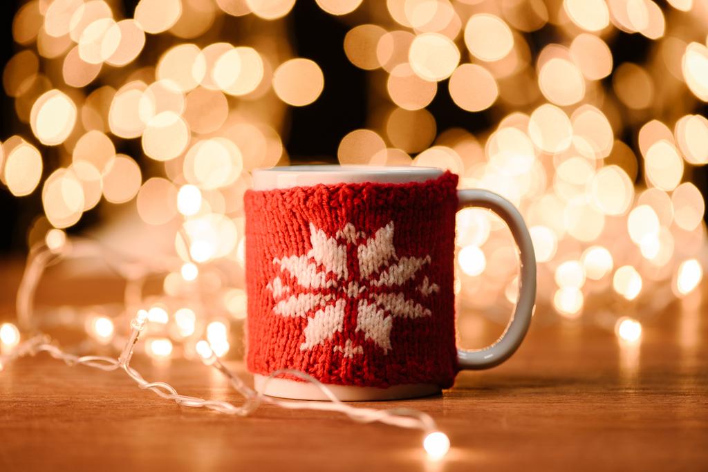 close up view of cup of hot drink and garland on wooden tabletop with bokeh lights background - Photo, Image