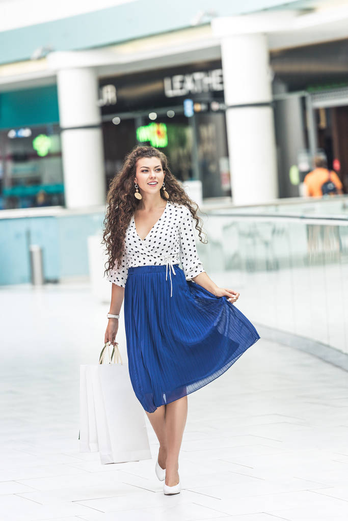 hermosa joven sonriente sosteniendo bolsas de papel y caminando en el centro comercial
 - Foto, Imagen