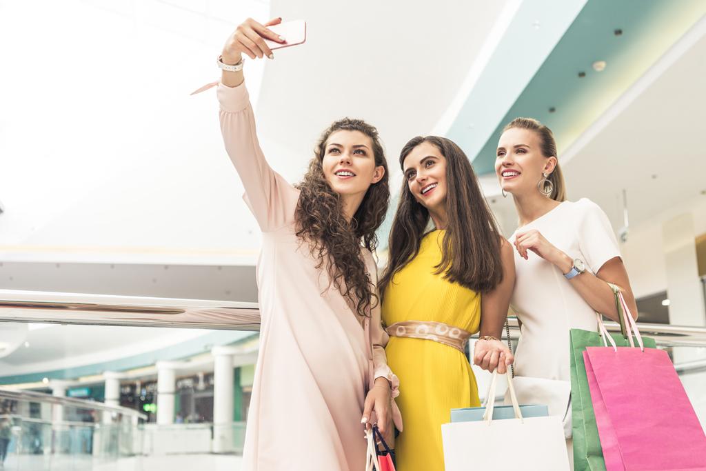 Blick auf glückliche, stylische Mädchen, die Papiertüten in der Hand halten und ein Selfie mit dem Smartphone im Einkaufszentrum machen - Foto, Bild