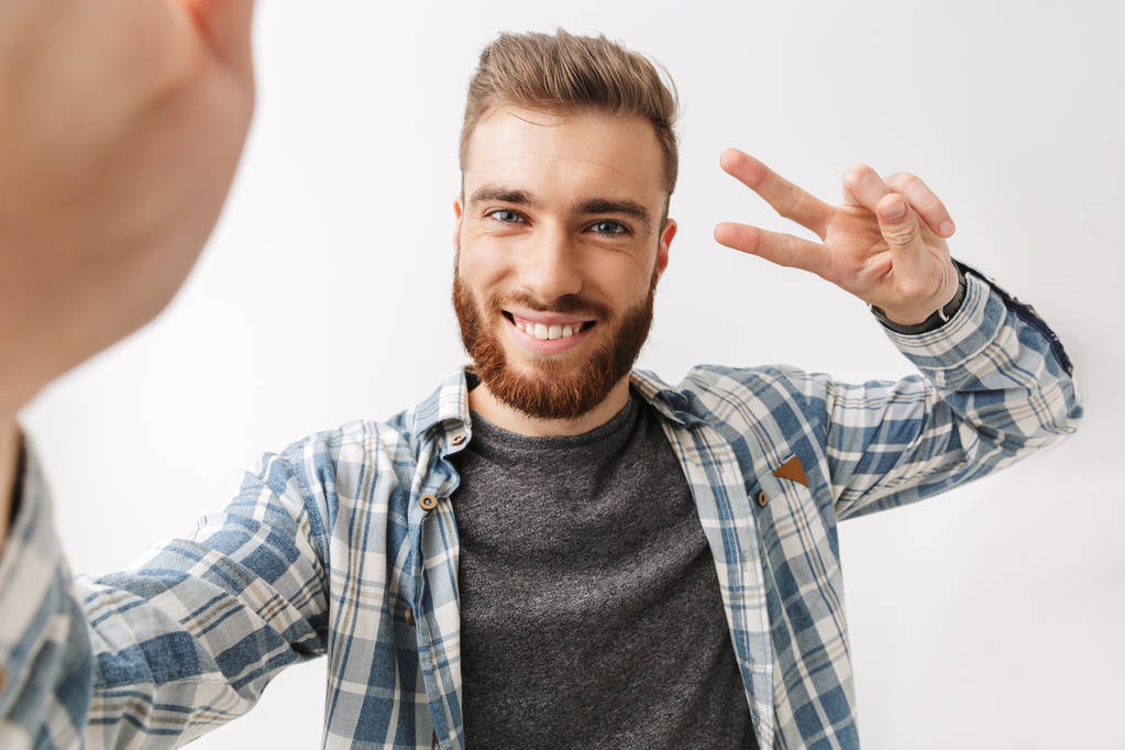 Portrait of a cheerful young bearded man standing isolated over white, taking a selfie, showing peace - Photo, Image