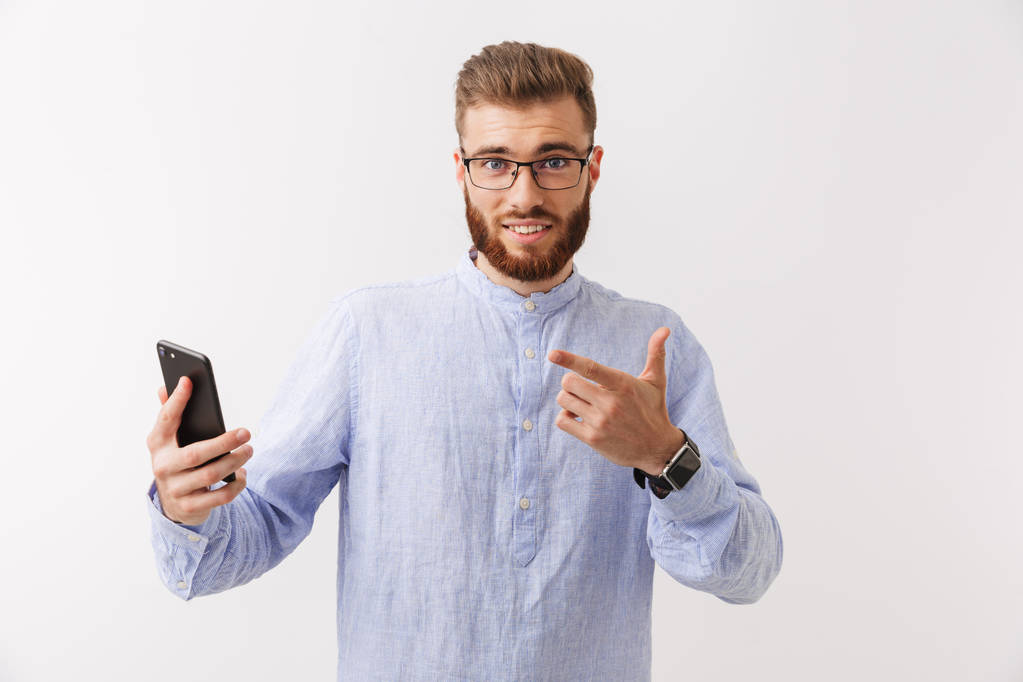 Портрет улыбающегося молодого бородатого человека в очках, стоящего изолированно над белым, держащего мобильный телефон
 - Фото, изображение