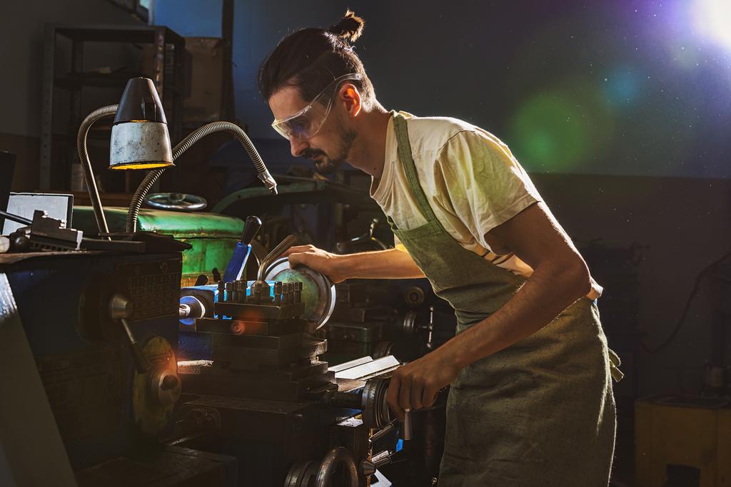 travailleur masculin concentré de fabrication dans le tablier et les lunettes de protection à l'aide de la machine-outil à l'usine
 - Photo, image