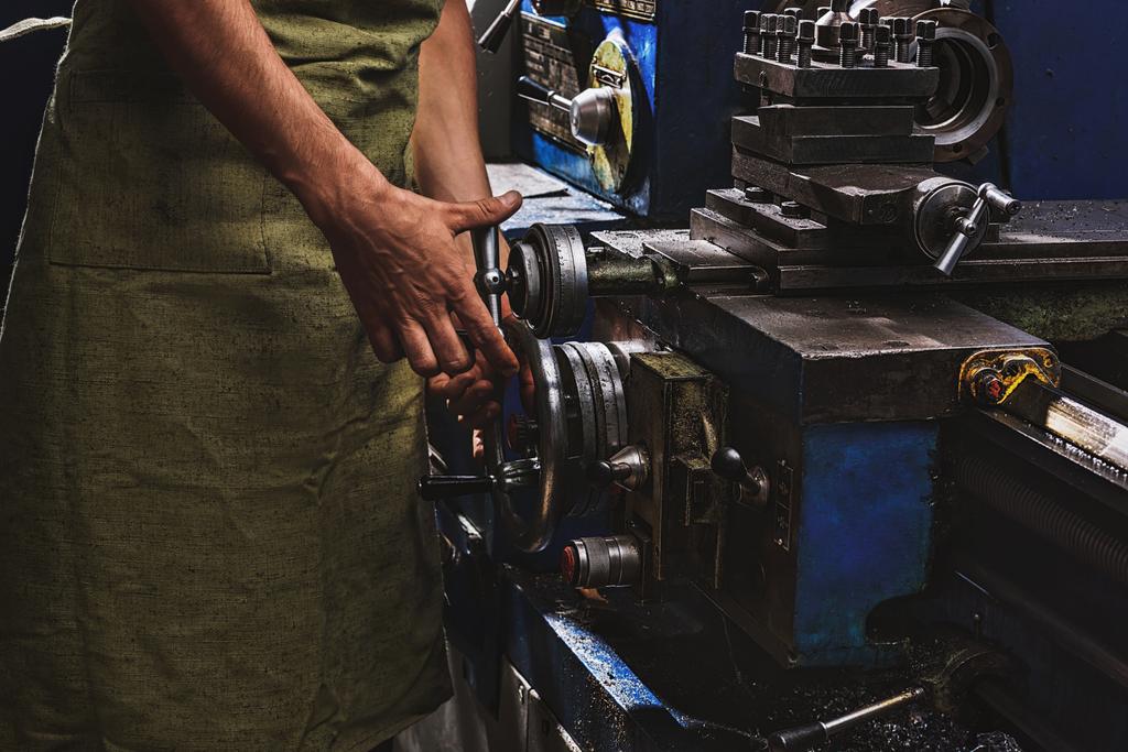 περικομμένη εικόνα της ανδρικής κατασκευής εργαζόμενος στην προστατευτική ποδιά χρησιμοποιώντας εργαλείο παραγωγής στο εργοστάσιο - Φωτογραφία, εικόνα