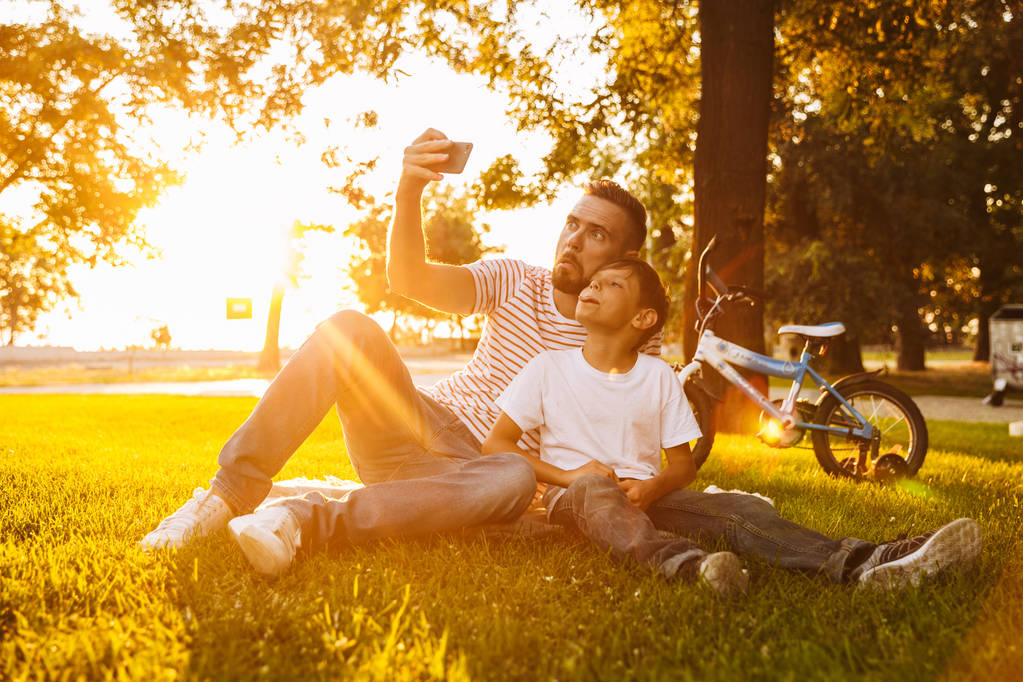 Alegre padre y su hijo se divierten juntos en el parque verde, tomando una selfie
 - Foto, Imagen