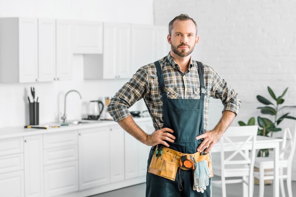 handsome plumber in overalls and tool belt looking at camera in kitchen - Photo, Image