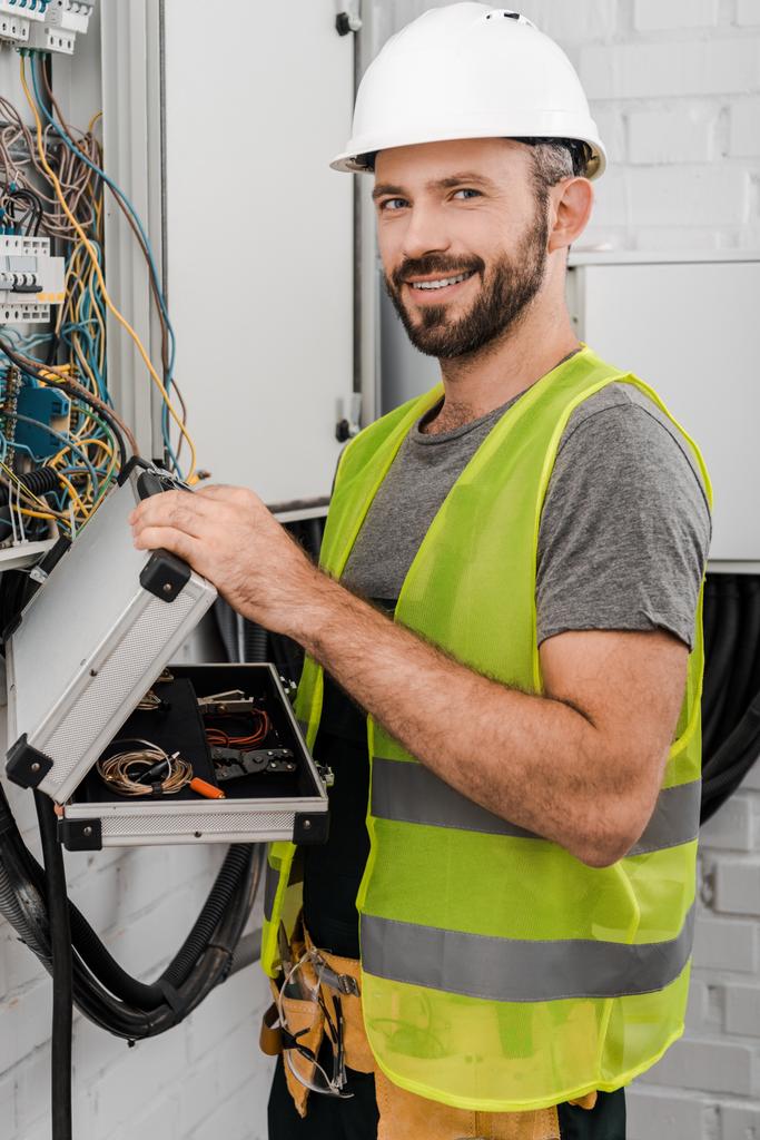 sonriente electricista guapo sosteniendo caja de herramientas cerca de la caja eléctrica en el pasillo y mirando a la cámara
 - Foto, imagen