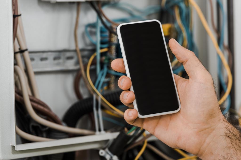 Oříznout obrázek elektrikář držení smartphone s prázdnou obrazovkou poblíž elektrické pole v chodbě - Fotografie, Obrázek