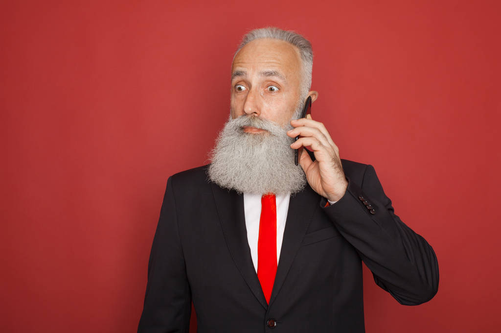 Πορτρέτο του έκπληκτος παλιά γενειοφόρος άνδρας με κοστούμι χρησιμοποιώντας κινητό τηλέφωνο που απομονώνονται σε κόκκινο φόντο - Φωτογραφία, εικόνα