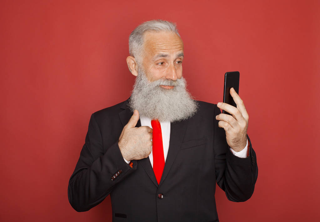 Studio záběr šťastný starší vousatý podnikatel s úsměvem při používání mobilního telefonu proti červené pozadí - Fotografie, Obrázek