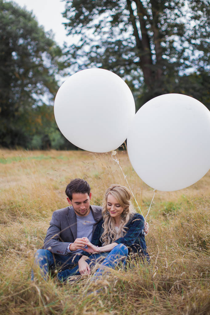 La joven pareja romántica feliz sentada en abrazo en la hierba al aire libre y sosteniendo los globos blancos en las manos y tomados de la mano
 - Foto, imagen